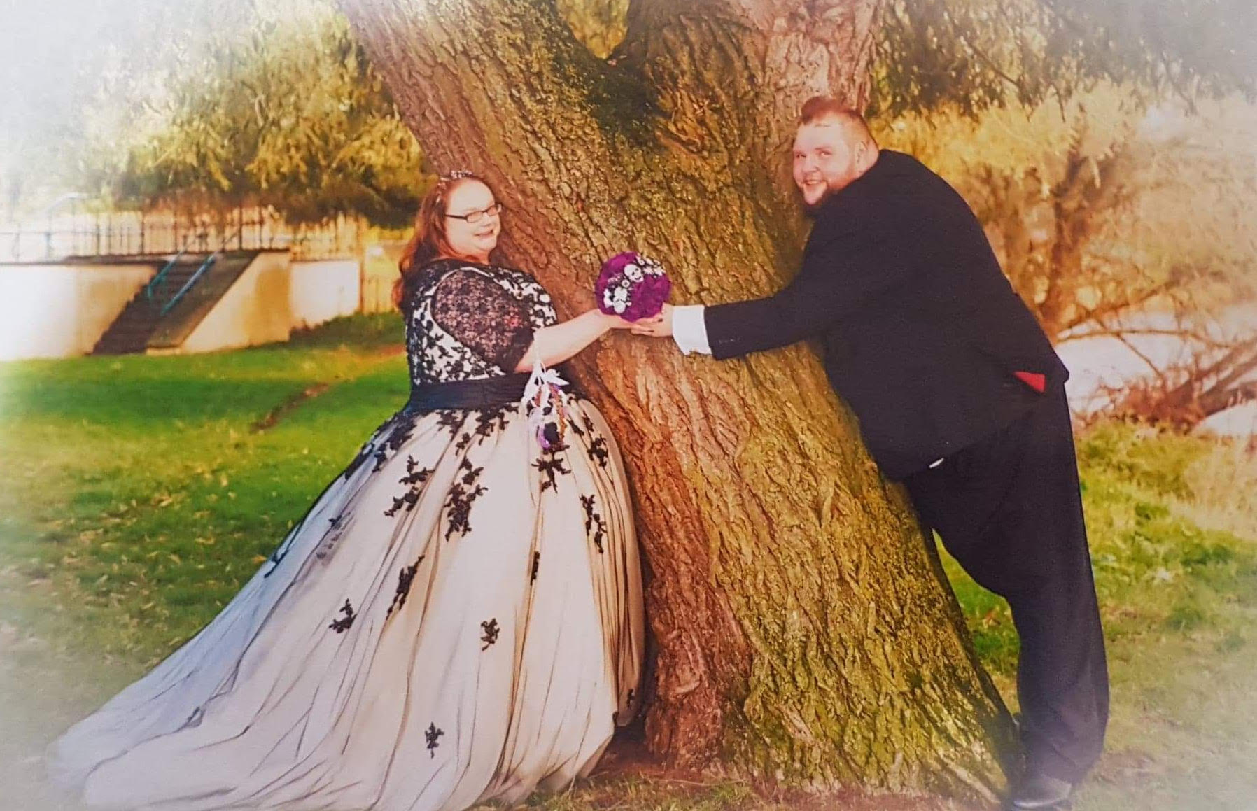 Kirsty wears a white and black wedding dress, with Chris in a black suit as they hold hands around a tree.
