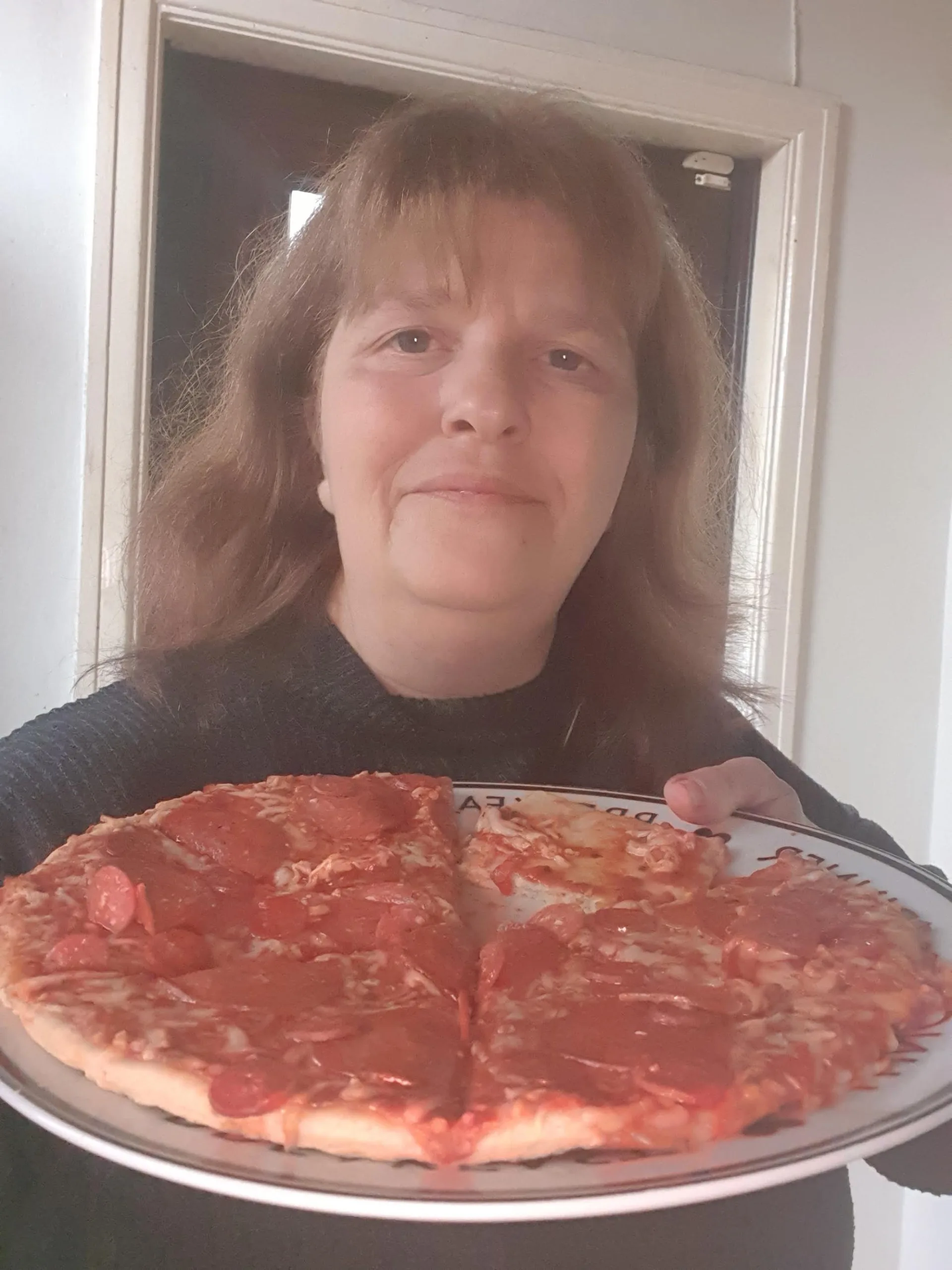 Janette is shown smiling while holding up a meat feast pizza.
