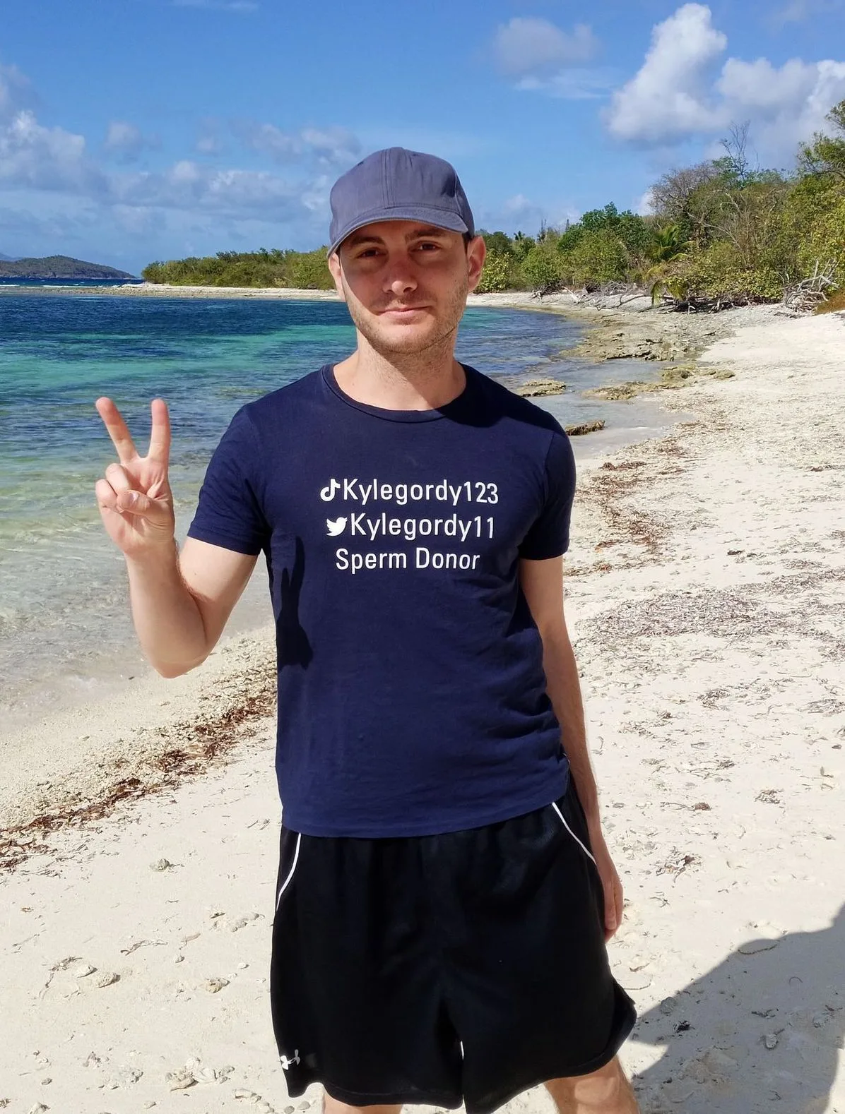 Kyle does a peace sign with his hand while on a beach.