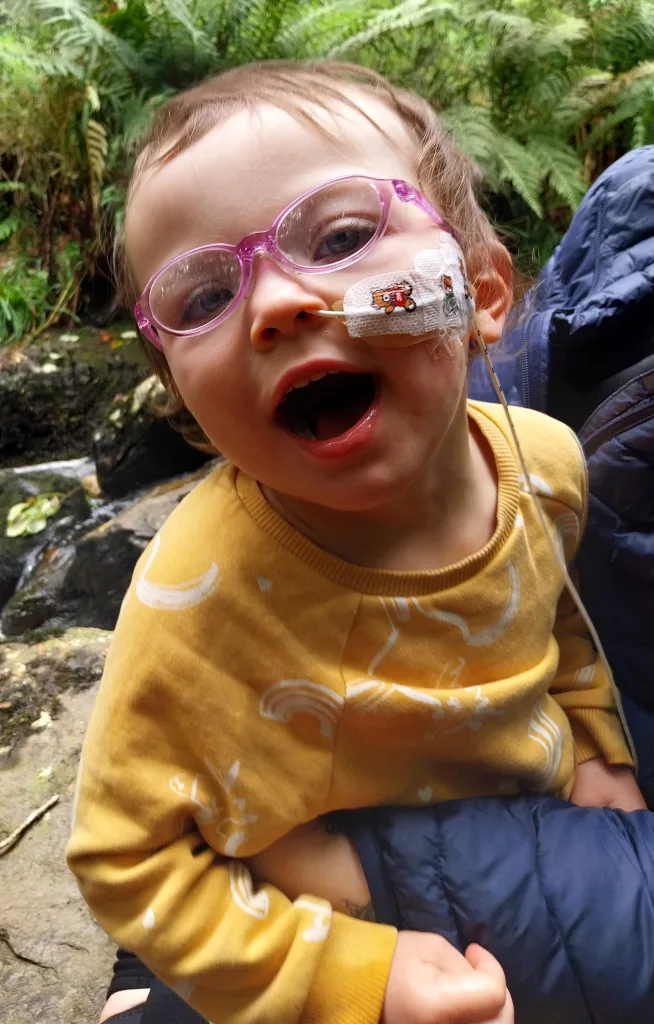 Iris-Rose smiles now with a tube in her nose, wearing pink glasses and a yellow jumper. 