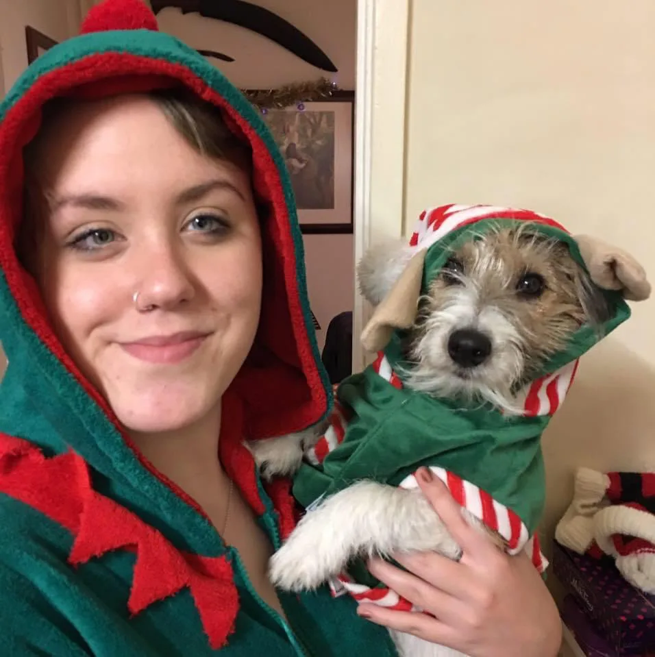 Charlotte smiles while wearing an elf costume and holding her dog who is also dressed in the same costume.