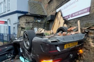Driver walks away after £60,000 Mercedes flips and crashes into London building - police seek occupants who fled scene as debris litters road and wall is destroyed.