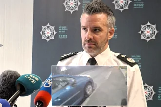A car bomb exploded yards from a police officer outside a Belfast station after a delivery driver was forced at gunpoint to transport the device, with families narrowly escaping injury.