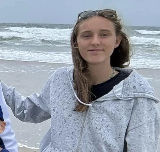 Anna Mellano standing on a beach in denim shorts and grey hoodie with sunglasses on head.