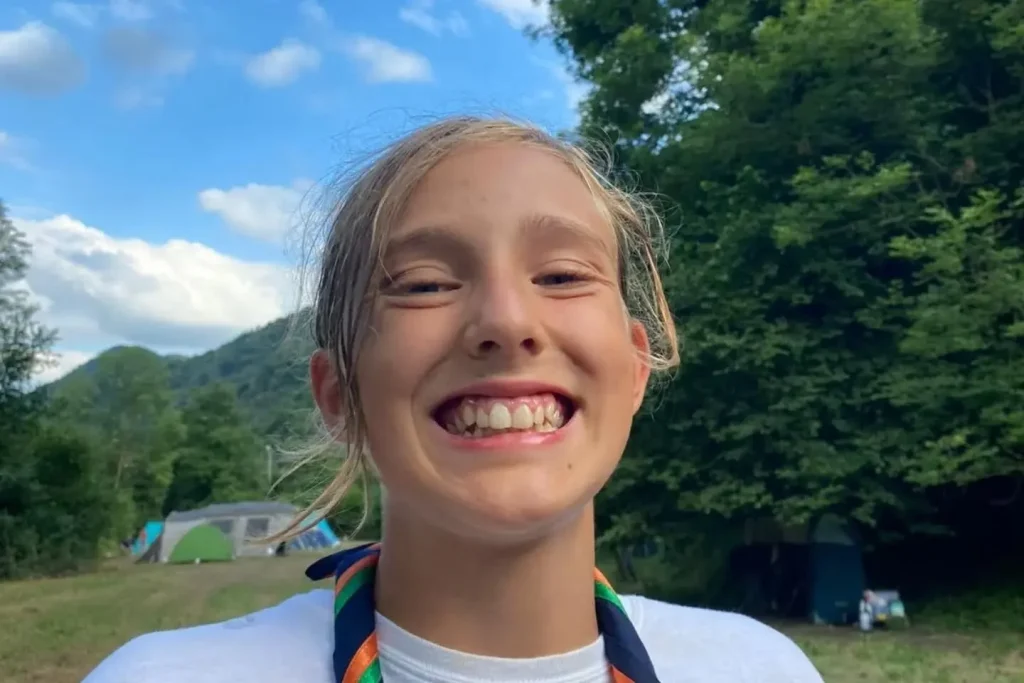 Young girl wearing white shirt with rainbow medal smiling outdoors with mountains behind.