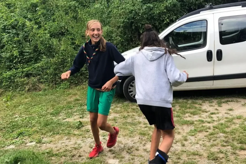 Two young people running together outdoors on grass with white van and trees in background.