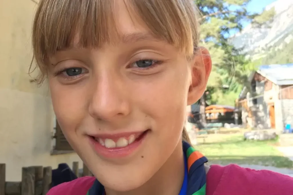 Young girl with light brown hair and fringe smiling at camera outdoors in sunshine.