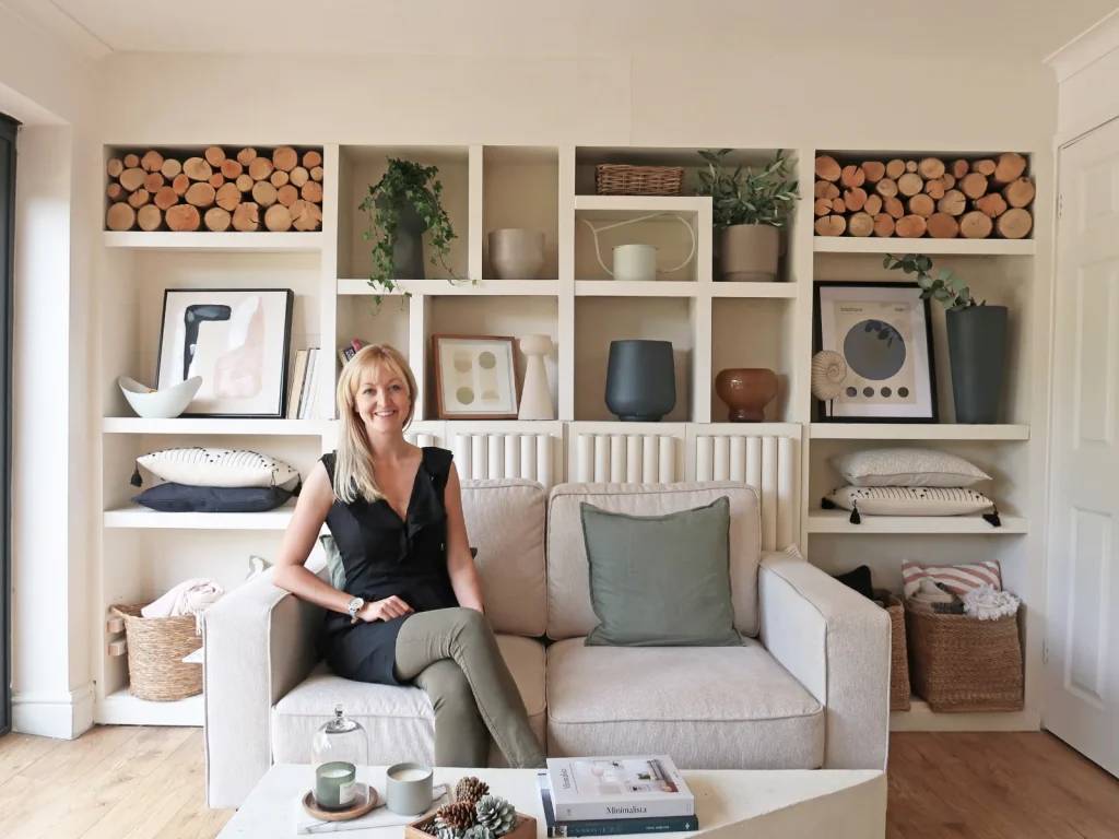 Woman sitting on beige sofa in front of white shelving unit displaying decorative items and firewood.