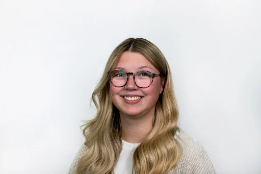 Young woman with glasses and long blonde hair, smiling at camera against white background.