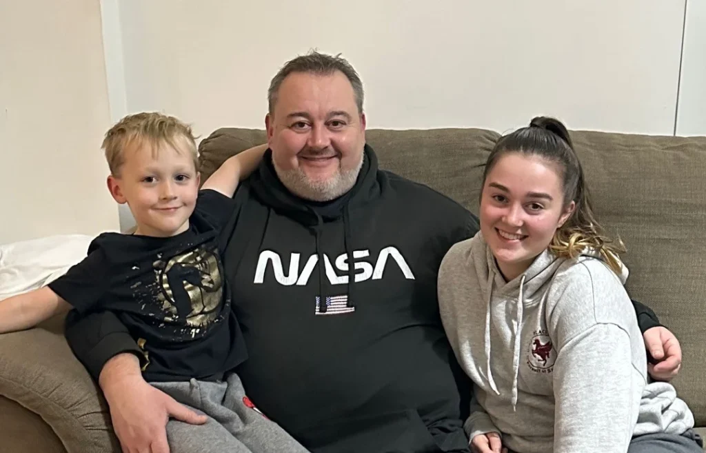 Man with two children sitting on beige sofa, all smiling at camera.