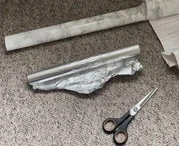Flat lay of rolled tin foil, self-adhesive vinyl, and scissors on grey carpet.