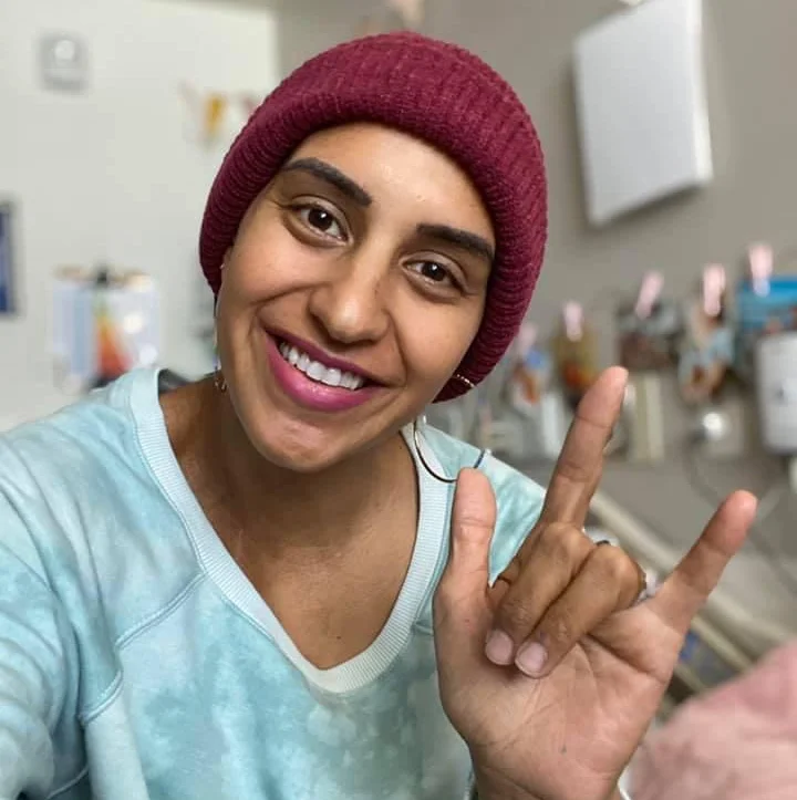 Woman in bathroom wearing maroon beanie and light blue shirt smiling at camera