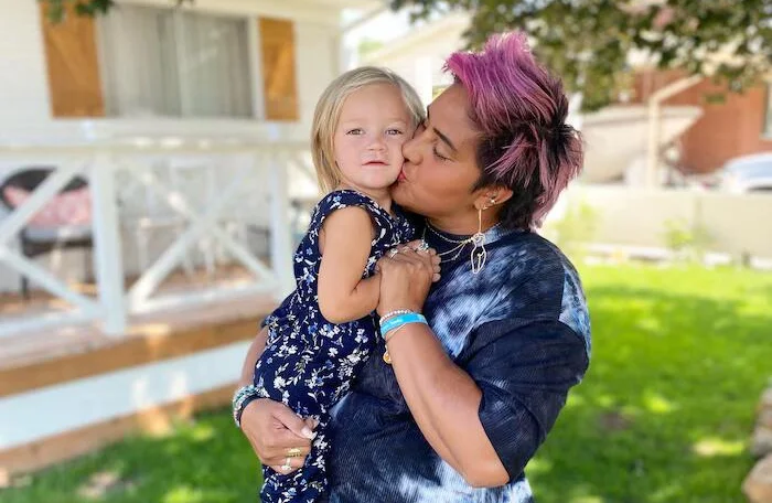 Woman with pink/purple hair holding toddler in floral outfit, outdoor daylight
