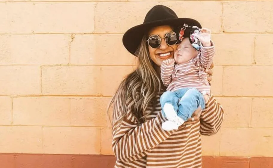 Woman wearing striped top and hat holding blonde infant in outdoor setting