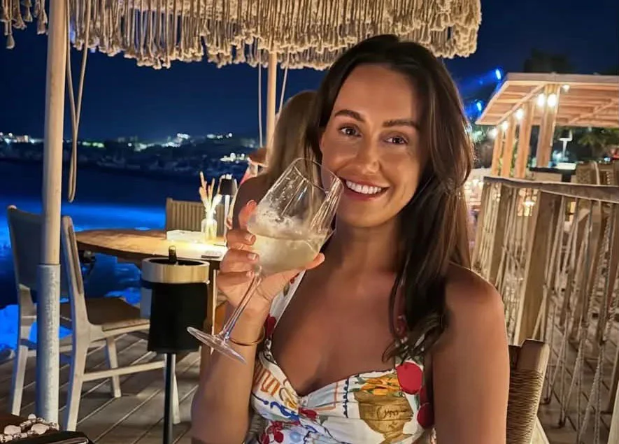 Woman with long dark hair holding white wine glass smiling at camera at waterfront restaurant
