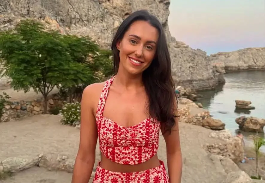 Woman with long dark hair smiling at camera in red patterned dress against coastal rocks and water