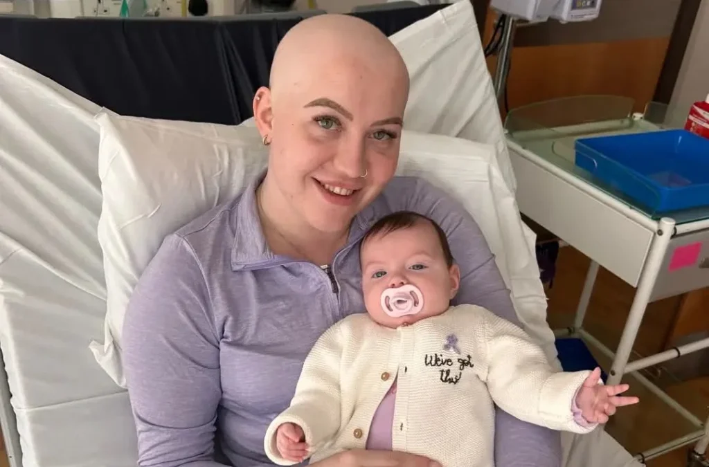 Woman with shaved head in hospital bed smiling whilst holding her young daughter Riven-Leigh.