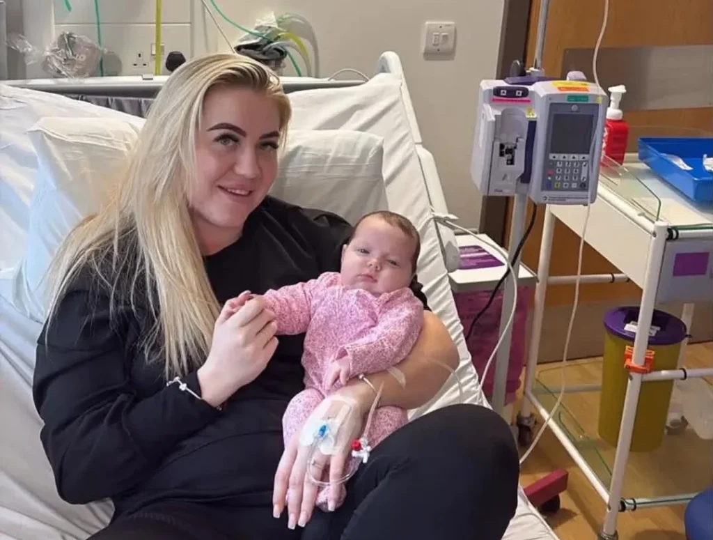 Woman in hospital gown with shaved head holding her infant daughter whilst sitting on the bed.