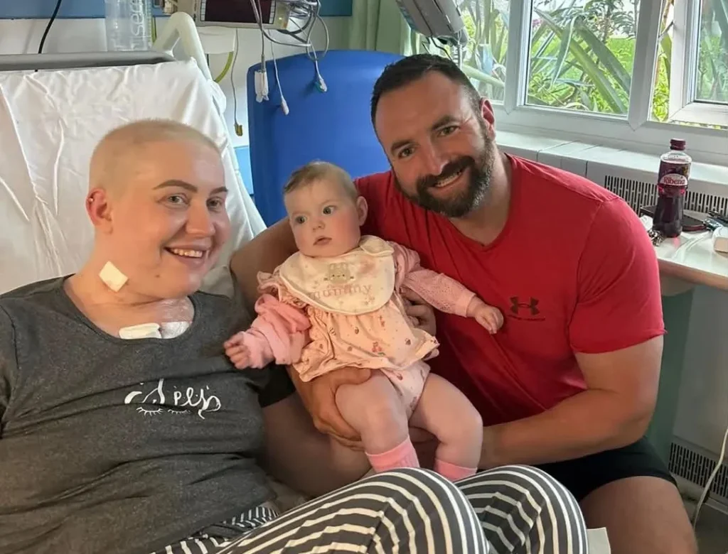 Woman in hospital bed with partner Richard and young daughter Riven-Leigh visiting.