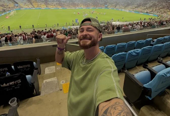 Fred Bruno at a large football stadium in a green shirt, making a peace sign to camera.