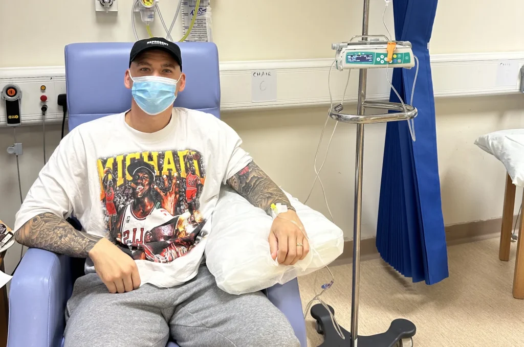 Matthew Spicer sitting in a blue chair during hospital treatment wearing a mask
