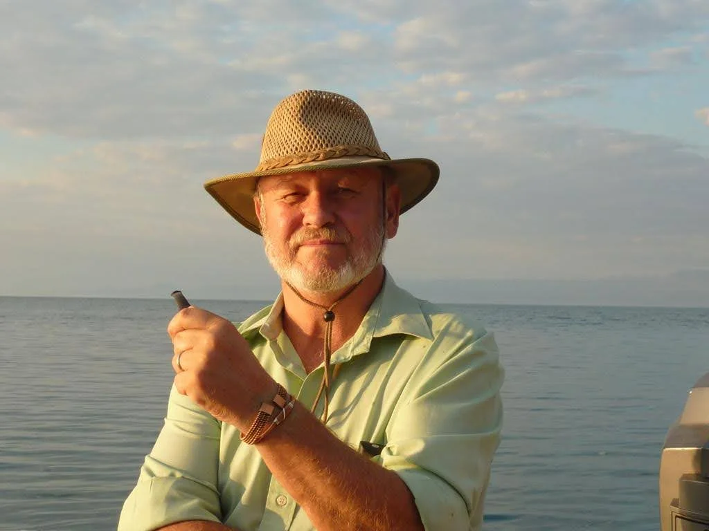 Simon, a man wearing a khaki hat and light shirt, posing outdoors by a calm sea.