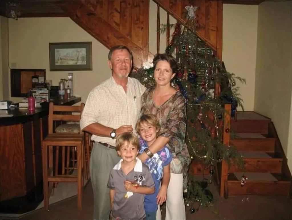 A man in khaki clothing with two young children standing indoors by a decorated Christmas tree.