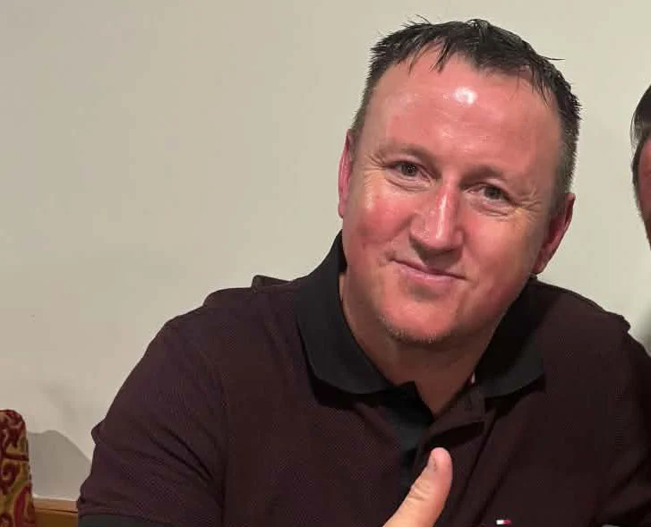 Edward Stokes, wearing a dark polo shirt, giving a thumbs up gesture while holding a drink.