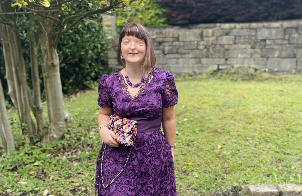 Woman with dark hair wearing purple patterned dress standing against garden stone wall.